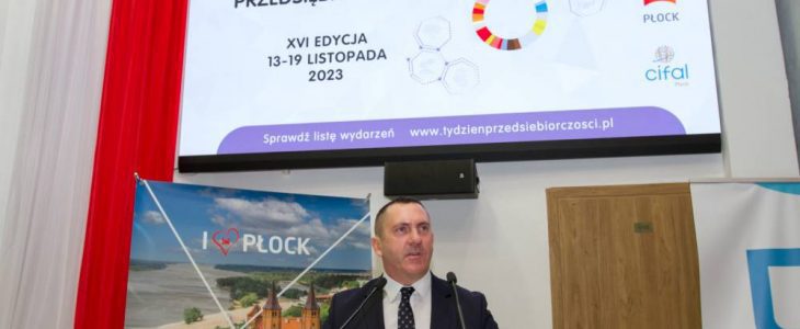 Wiceprezydent Artur Zieliński przemawia do mikrofonów na mównicy. W tle plakat "I love Płock z widokiem z tumskiego wzgórza, a wysoko nad głową na wielkim ekranie wyświetlany jest slajd tytułowy rozpoczynający Światowy Tydzień Przedsiębiorczości w ubiegłym roku.